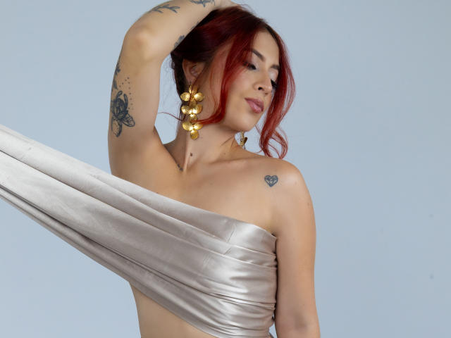 MiaaRouge with red hair, gold earrings, and tattoos, wrapping a silver fabric around her tits, looking down with a slight smile, arm raised. Sheer skin, confident pose.