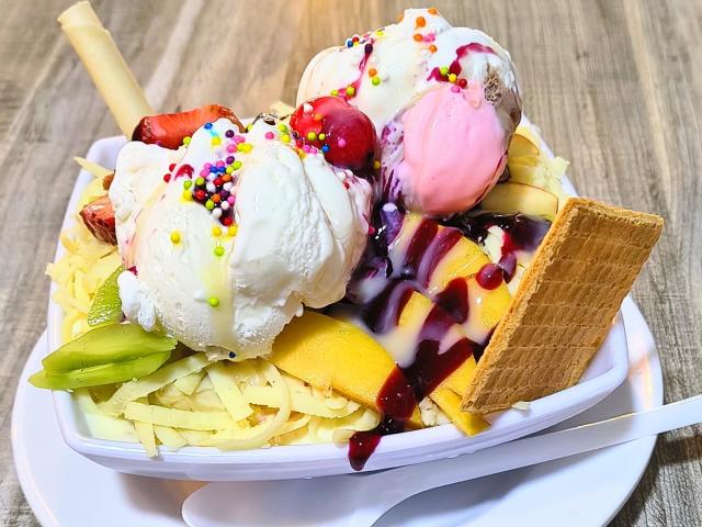 A creamy, colorful ice cream sundae with two scoops, sprinkles, cherry, banana, waffle, and strawberry, on a white plate with a spoon, featuring a juicy green apple slice.