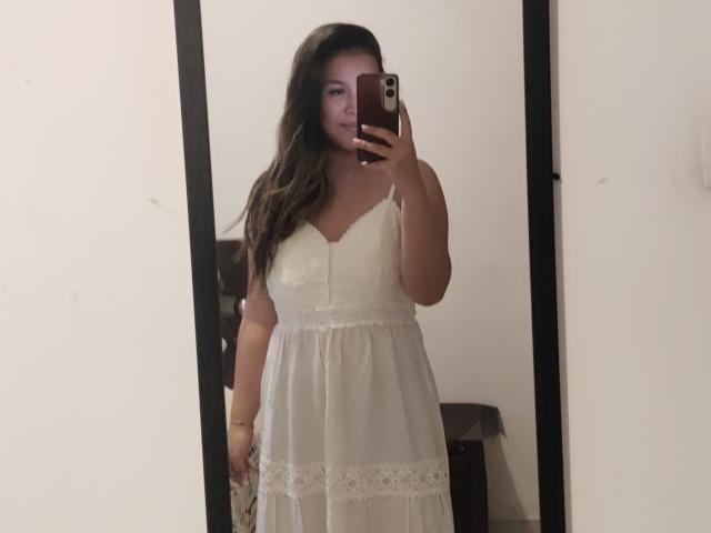 LiaDiane with long brown hair wearing a white lace dress, standing in front of a mirror, holding a phone, showing her tits and a subtle ass.
