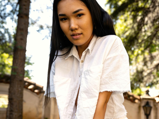DivineMadness, Asian woman with long black hair, wearing a white button-up shirt, standing outdoors, looking slightly down, sunlight filtering through trees, soft smile, casual, natural light.