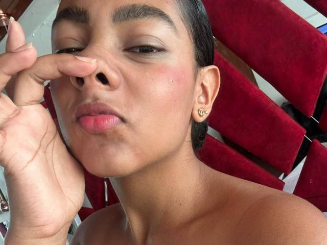 NatalyBlonder's face, nose pinched between fingers, brown skin, short black hair, small gold hoop earring, pinkish lips, slight blush, bare shoulders, red velvet chair in background.