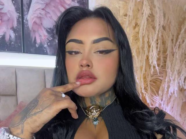 LairaHearvy with black hair, tattooed neck, and thick eyebrows, biting finger with gold ring, wearing black top with rose pendant, and pink lipstick.
