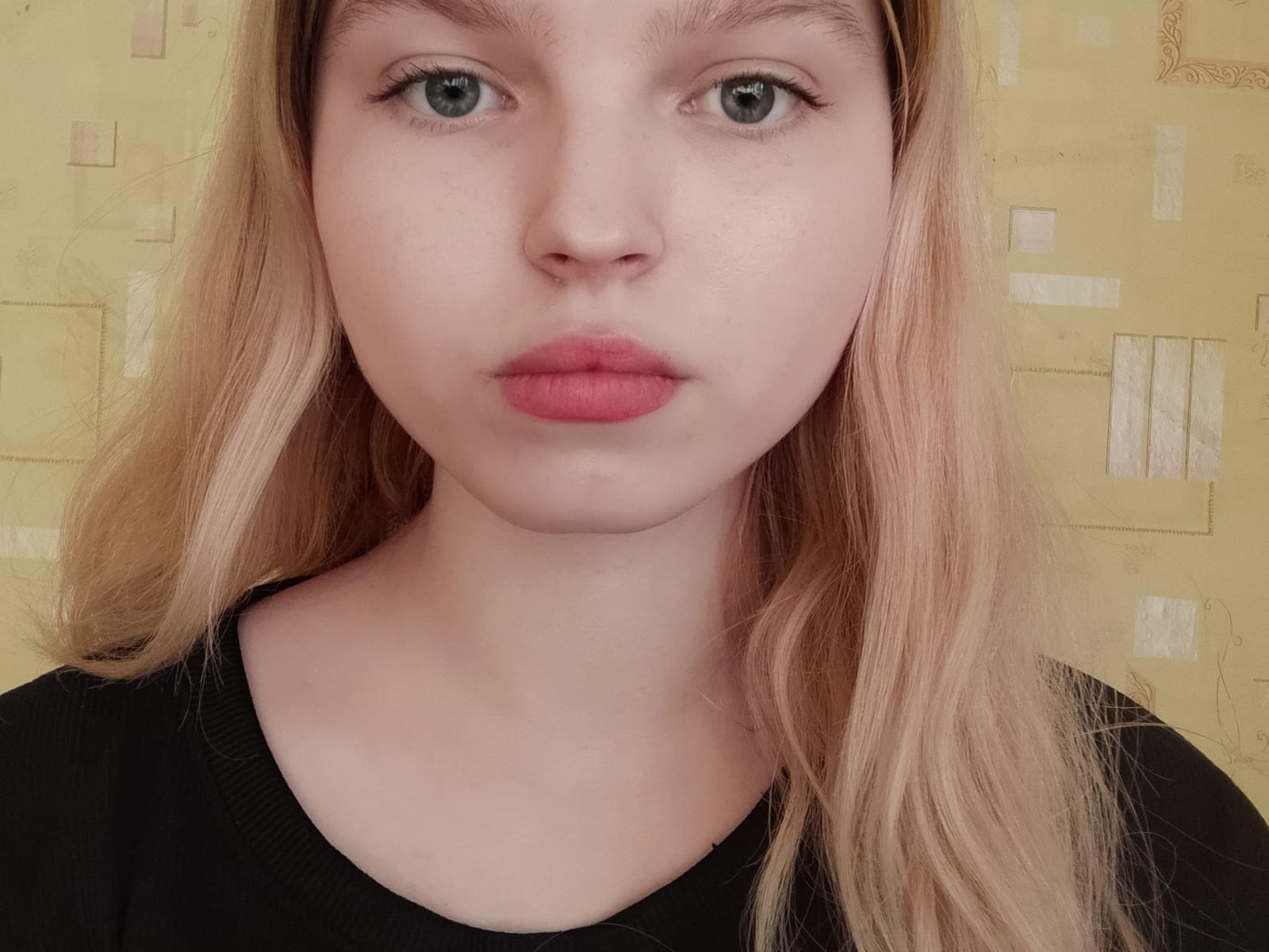 MaximaLasley hat hellgrüne Augen, rosa-rote Lippen und glattes blondes Haar und trägt ein schwarzes Hemd. Ihre helle Haut und ein leichter Rouge-Ton sind sichtbar. Sie wirkt ernst..