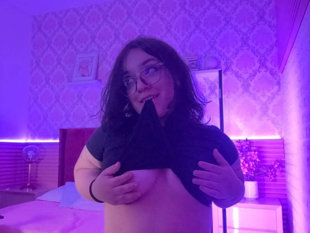 NiniWillow with glasses, medium brown curly hair, lifting black shirt, exposing bare tits, biting black triangle, purple bedroom lighting, smiling.