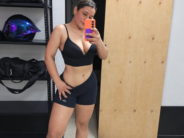 EmaFoxy with dark hair in a black sports bra and black shorts, showing her tits and ass, taking a mirror selfie with an orange phone, hand on her crotch.