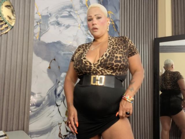 Gianella Cox, con cabello blanco corto, vestida con una blusa de estampado de leopardo y una falda negra con un cinturón grande, levantando la falda, mostrando sus glúteos y mirando el espejo..