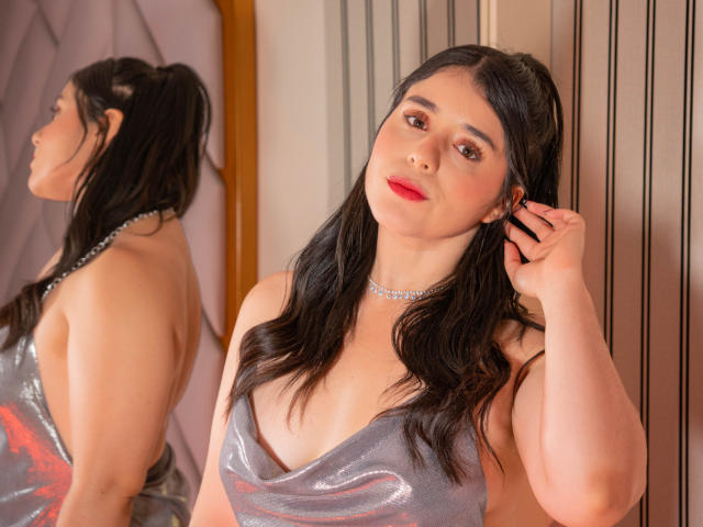 VickyMegan with black hair, red lipstick, and a shiny silver dress, her hand in her hair, wearing a choker, her reflection in the mirror showing her back.