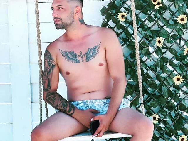 TommyDum, torse nu et couvert de tatouages, est assis sur un banc blanc, vêtu d'un short bleu et blanc, tenant un téléphone noir, et protégeant ses parties génitales, devant un mur blanc orné de tournesols..