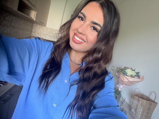 Elefteria, con i capelli castano ondulati, indossa una camicia blu, sorride mostrando i denti bianchi, ha gli occhi marroni e porta una collana d'argento. Il suo seno non è visibile. Sta scattando un selfie..