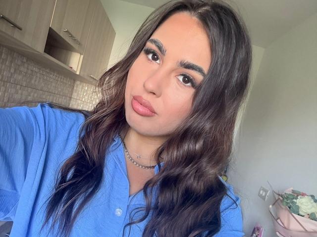 ElefteriaBlis mit welligem, dunkelbraunem Haar, einem blauen Hemd und rosafarbenen Lippen, trägt eine silberne Halskette, blickt mit braunen Augen in die Kamera, hat dezentes Make-up und einen leichten Schmollmund..