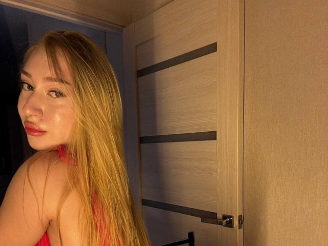 JoetteShigeta with long blonde hair, red lips, and a slight smile, showing her bare shoulders and pink top, standing near a wooden door. Her tits are visible.