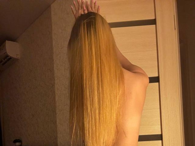 Topless JoetteShigeta with long blonde hair, arms raised, back to camera, hands on door, warm indoor lighting, wooden door, beige wall, air conditioning unit on left.