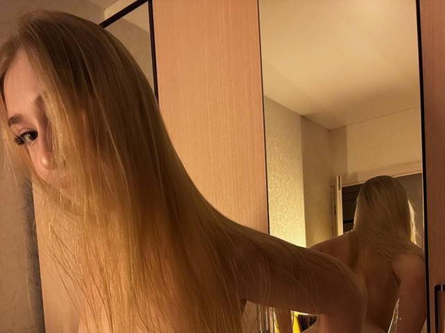 JoetteShigeta with long blonde hair, looking over her shoulder, reflection in mirror shows bare back and butt, soft warm lighting, gentle expression, subtle makeup, in a cozy room.