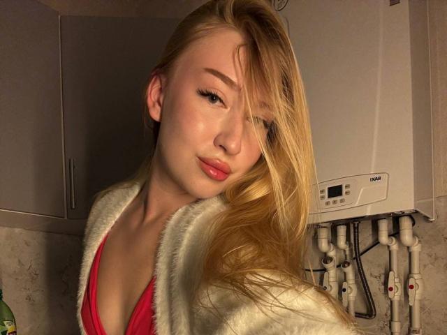 JoetteShigeta with blonde hair, red top, white fluffy jacket, slightly parted lips, soft smile, small tits, fair skin, light makeup, standing in a dimly lit kitchen.
