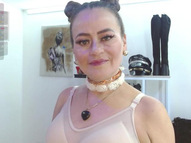 Maghentaa with two buns in her hair, pink lipstick, white lace choker, pink mesh top, black heart necklace, smiling, standing, wearing earrings, black boots in background, white wall.