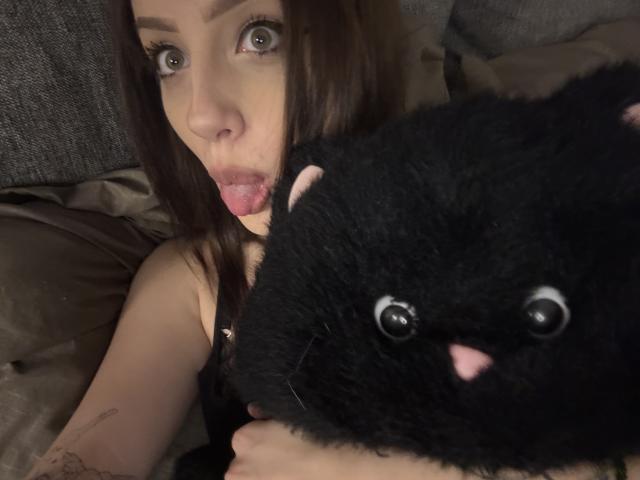 SimonneBrustkern with wide eyes and tongue out, holding a black stuffed cat with pink nose and white eyes, in a dimly lit, cozy setting.