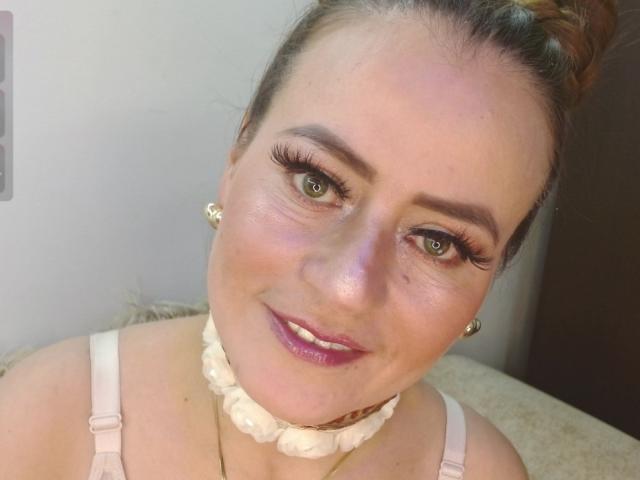 Maghentaa has light skin, green eyes, and thick eyebrows with long eyelashes. She's wearing a white ruffled choker and pink bra, smiling with pink lipstick.