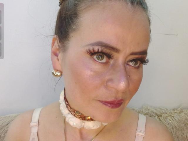 Maghentaa with brown hair in a bun, wearing a white bra, has green eyes, long eyelashes, and a brown choker with white flowers, and red lipstick, smiling slightly.