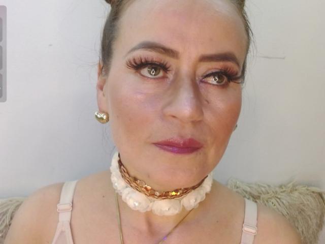 Maghentaa has fair skin, green eyes, and pink lipstick. She's wearing a white floral choker, pink bra, and gold earrings. Her hair is in a bun, looking serious.