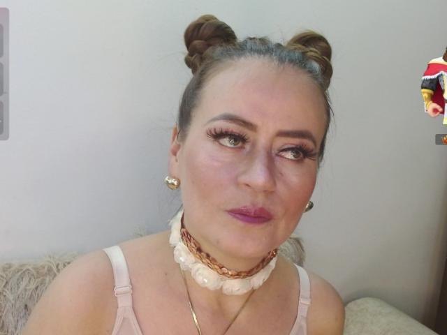 Maghentaa has brown double buns, thick eyelashes, purple lipstick, white floral choker, gold earrings, and a pink bra, looking slightly to the side with a neutral expression.