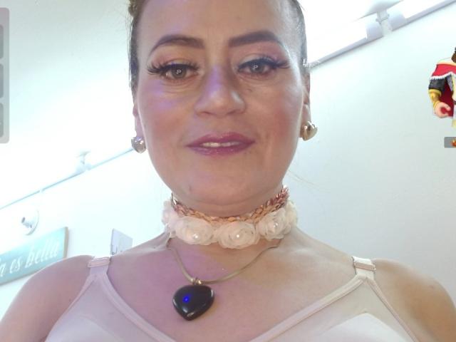 Maghentaa with light skin, brown hair in a bun, wearing a pink top, heart necklace, and floral choker, smiling at the camera, showing off her tits.