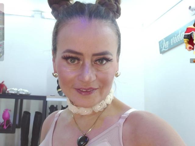 Maghentaa a des chignons bruns, de longs cils, du rouge à lèvres rose et un collier de roses blanches. Elle porte un haut rose, affiche un sourire adorable et une chaîne de pendentifs noirs..