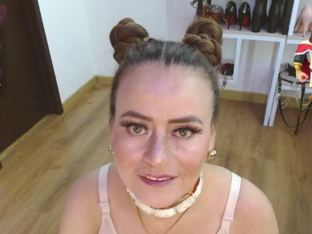 Maghentaa hat zwei Duttfrisuren, grüne Augen, dichte Wimpern, pinken Lippenstift und eine weiße Perlenkette. Sie trägt ein pinkes Oberteil, und ihre Brüste sind leicht sichtbar..