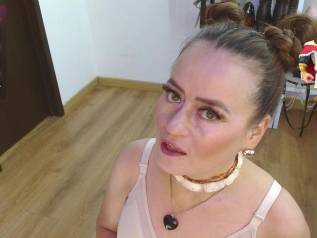 Maghentaa avec des chignons bruns, du rouge à lèvres rose, des yeux verts, un haut en dentelle blanche, un collier ras du cou, un collier de perles noires, un sol en bois et un miroir en arrière-plan. Les seins sont visibles à travers le haut en dentelle..