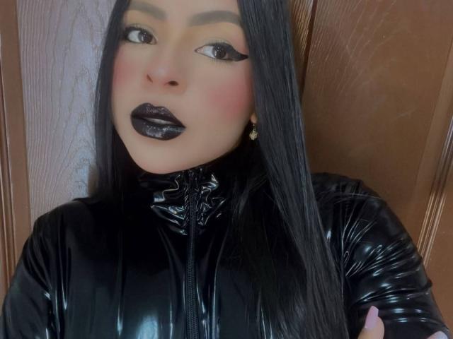 LindaFrancoo, vêtue d'une combinaison en latex noir, avec un rouge à lèvres noir brillant, de longs cheveux noirs et un eye-liner foncé, regardant la caméra, avec un fond en bois, portant de petites boucles d'oreilles dorées. Les seins ne sont pas visibles..