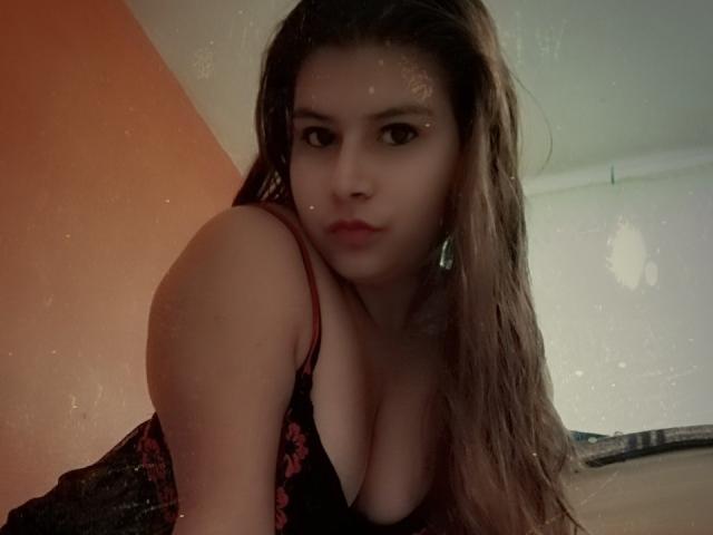 AllisonZIA with wet brown hair, red floral dress, showing cleavage, leaning forward, looking at camera with slightly parted red lips. Her tits are visible, and she's in a dim room.