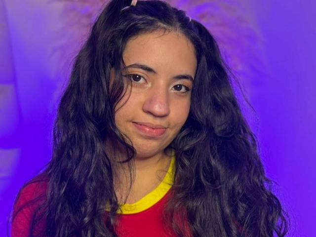 KrissChang has long, wavy black hair, a cute face, and a small smile. She's wearing a red shirt with yellow trim. Her skin is light brown.