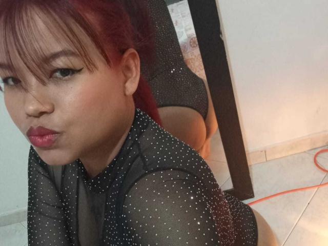 MelanieDiiaz with red hair, black sheer polka dot top, pouty red lips, kneeling, showing her ass in black mesh panties, looking at camera.