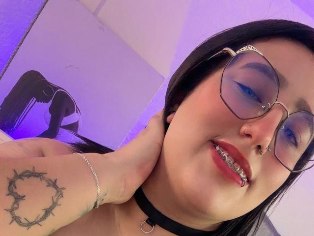 Natalie Harris, avec ses cheveux noirs, ses lunettes, son fard à paupières bleu, son piercing au nez et son rouge à lèvres rouge, mordant sa lèvre inférieure, portant un collier noir et touchant son cou. Elle a un tatouage sur sa main droite..
