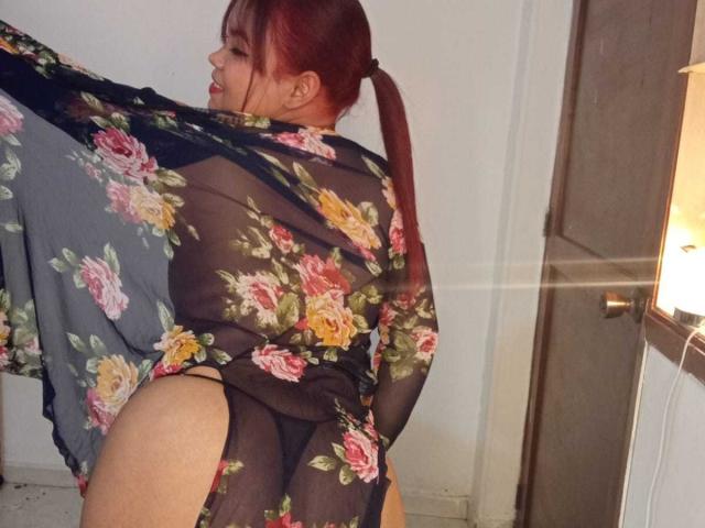 MelanieDiiaz with red pigtails, wearing a sheer floral robe, exposing her black thong and round ass. She's lifting the robe, showing her back and a side view.