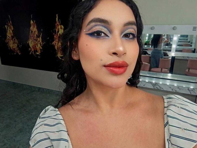 AdalinaVera has dark curly hair, blue eye makeup, red lipstick, small moles on her face, and wears a striped white top. She's standing in a mirror-lined room.