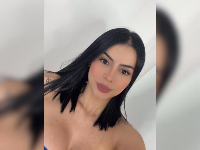 Isabella James heeft zwart, schouderlang haar, donkere wenkbrauwen, roze lippenstift en een subtiele glimlach. Ze draagt geen top en haar kleine, onbedekte borsten zijn zichtbaar. Haar reflectie is lichtjes wazig op de voorgrond..