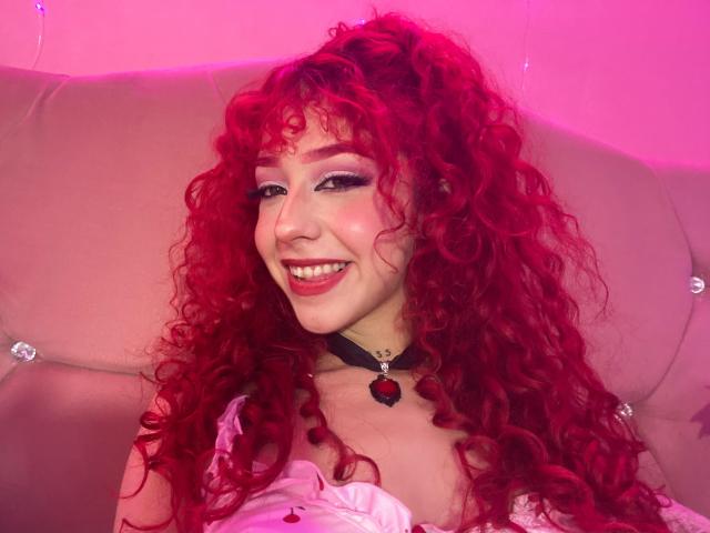 GaiaDoll with bright red curly hair, smiling, wearing a black choker with a red gem, pink makeup, and a white dress, sitting against a pink upholstered headboard.