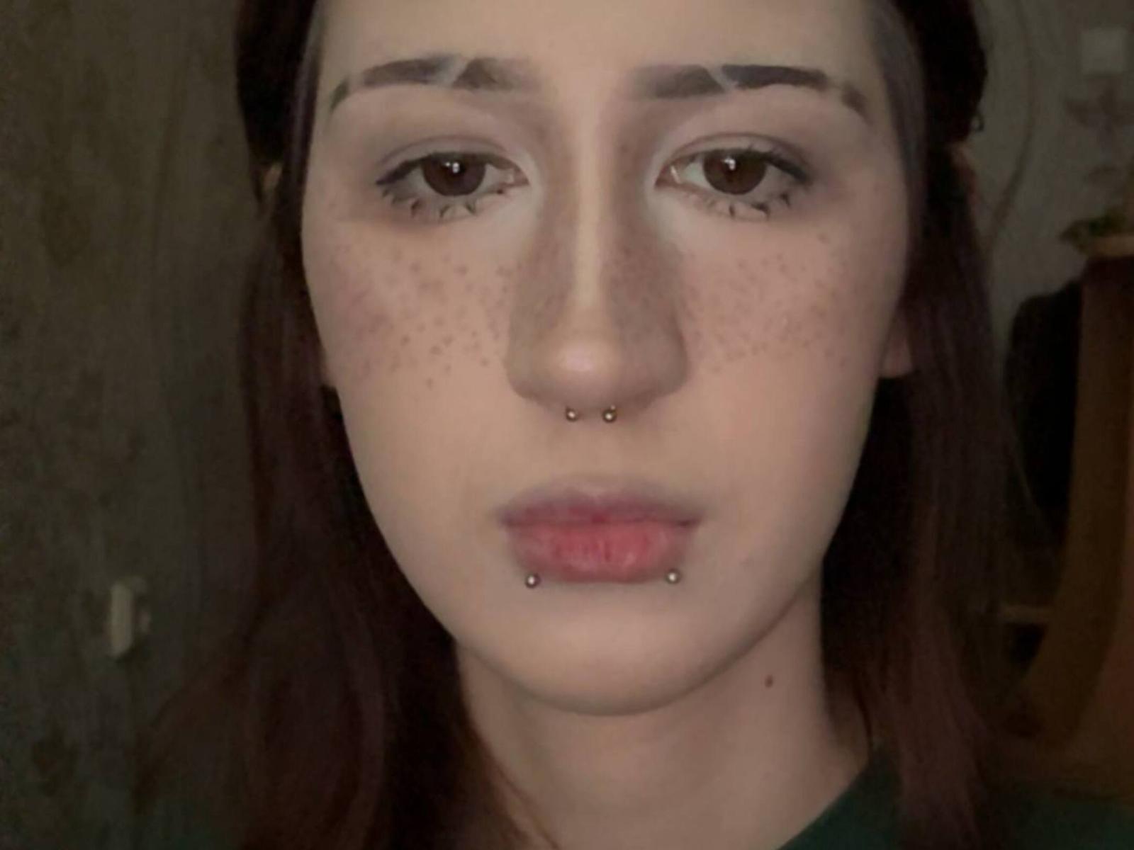 Jeanene Smolik ha delle lentiggini sul viso, occhi marroni, sopracciglia scure e dei piercing al naso e al labbro inferiore. Indossa una maglietta verde e ha i capelli castani..