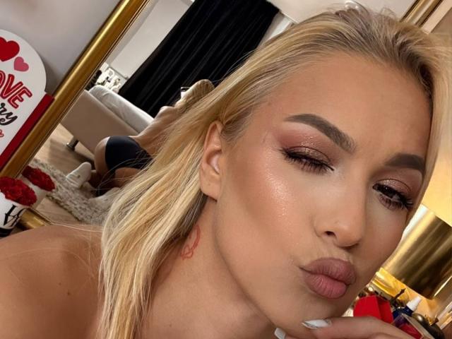 RebelRoss with pouty lips, blonde hair, and shimmering makeup, showing off her tits in the mirror. She's got pink eyeshadow and glossy lips, with a tattoo on her neck.