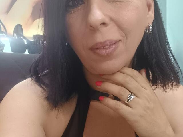 LauraSin has dark black hair, pink lipstick, red nails, and a silver ring. She's winking with her hand on her neck, showing off her tits. Her skin is tan.