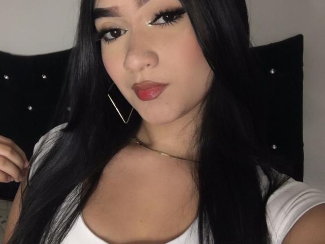 MiaScarleth69 a de longs cheveux noirs, des lèvres rouges pulpeuses, et porte un haut blanc avec des boucles d'oreilles dorées. Elle a un nez mignon et un collier en or..