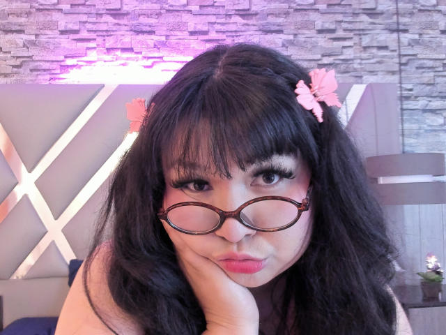 LillyMayer with long black hair, pink butterfly hair clips, big eyelashes, and round glasses, pouting with her hand on her cheek, showing off her cute ass.
