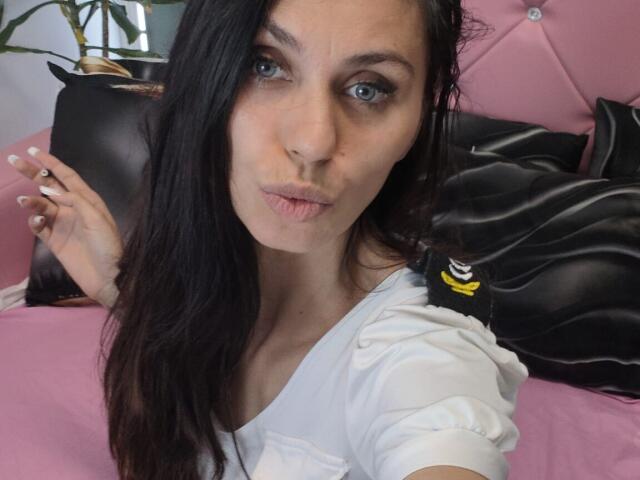 DanaWaw with pouty lips, blue eyes, and long black hair, wearing a white shirt, holding a cigarette, pink and black background. Boobs are not visible.
