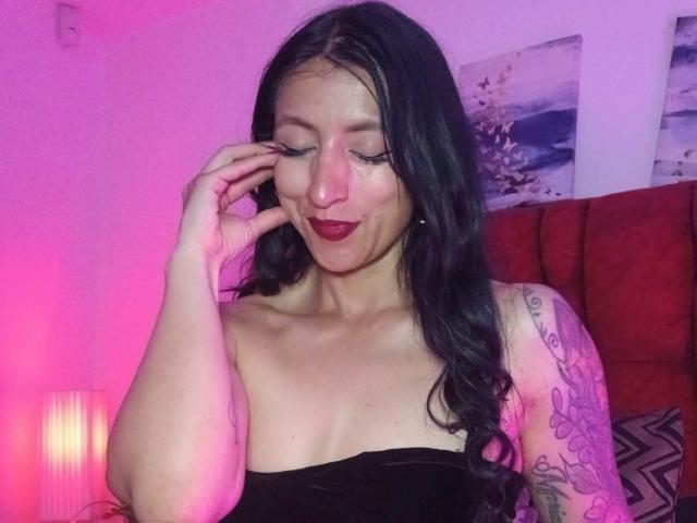 ZarayFeet ha lunghi capelli neri, rossetto rosso e un tatuaggio sul braccio destro. Sorride con gli occhi chiusi, toccandosi la guancia, e indossa un top nero senza spalline..