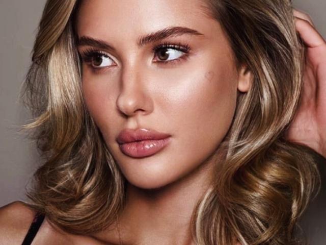 SensualMila has blonde, wavy hair, full, shiny lips, and brown eyes. Her skin is smooth with a slight glow, and she's looking to the side with a serious expression.