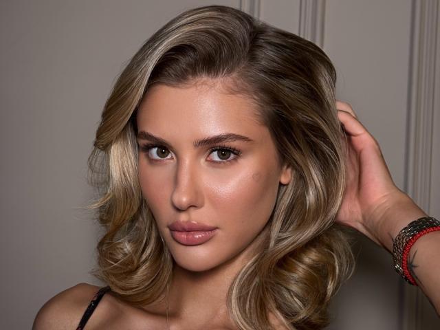 SensualMila met golvend blond haar, bruine ogen en glanzende roze lippen, draagt een zwarte top met bandjes, heeft haar hand in haar haar, een rode en zwarte armband en een subtiele, sexy grijns..