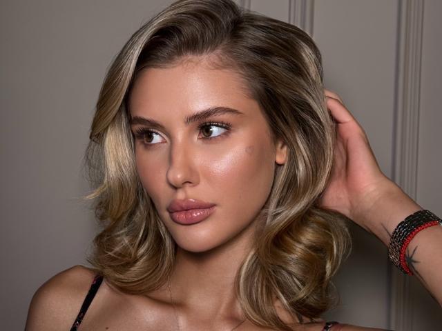 SensualMila met glanzend blond haar, glanzende roze lippen en een subtiele bruine huid, kijkt opzij met een hand in haar haar. Ze draagt een zwarte band, een kralenarmband..