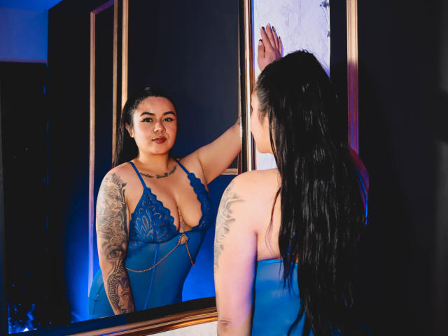 EveWillis with wet black hair, tattoos on arms, wearing a blue lace-up bodysuit, staring at herself in a mirror, touching it with her right hand. Boobs visible.