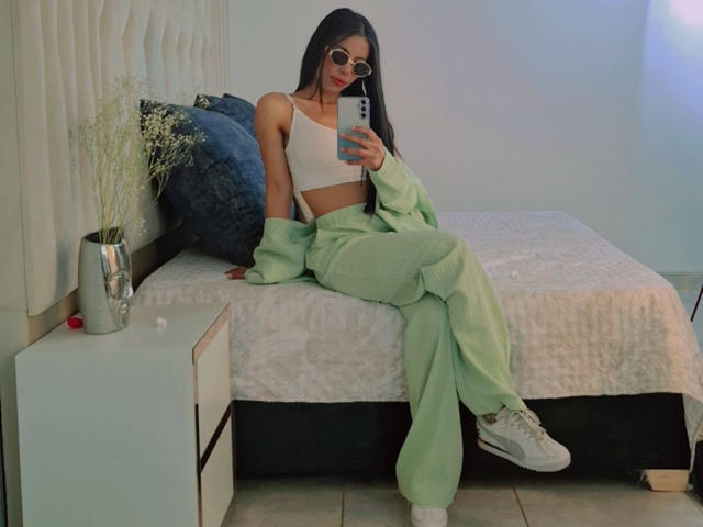 Laura Jackson, con senos pequeños, pantalones verdes, top corto blanco y chaqueta verde, sentada en la cama, sosteniendo un teléfono, con zapatillas blancas, cabello largo y oscuro y gafas, en un dormitorio..