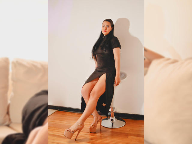ElizabethDupont with long black hair, wearing a sheer black dress with a thigh slit, sitting on a stool, showing her ass, in nude platform heels, wooden floor.